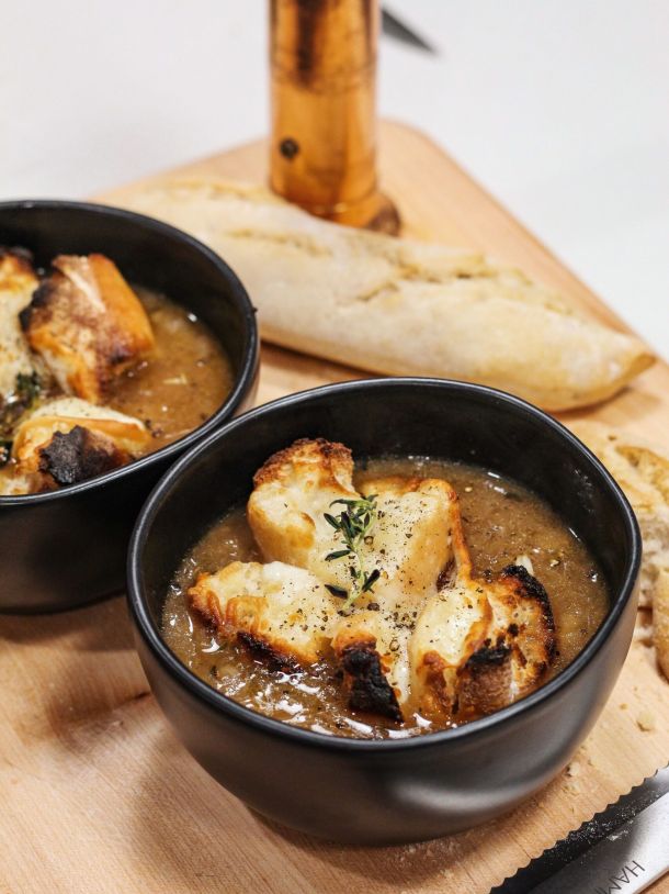 Two black bowls of creamy soup topped with croutons and herbs on a wooden board, with a torn baguette in the background.