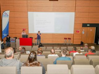 Speaker at a red lectern presenting to an audience in a conference hall, with a large projected slide on a wood-paneled wall behind them.