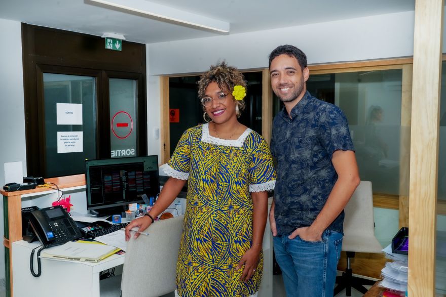Two coworkers pose for a photo in an office reception area, the woman in a bright yellow patterned dress with a yellow flower in her hair and the man in a blue patterned shirt smiling beside her.