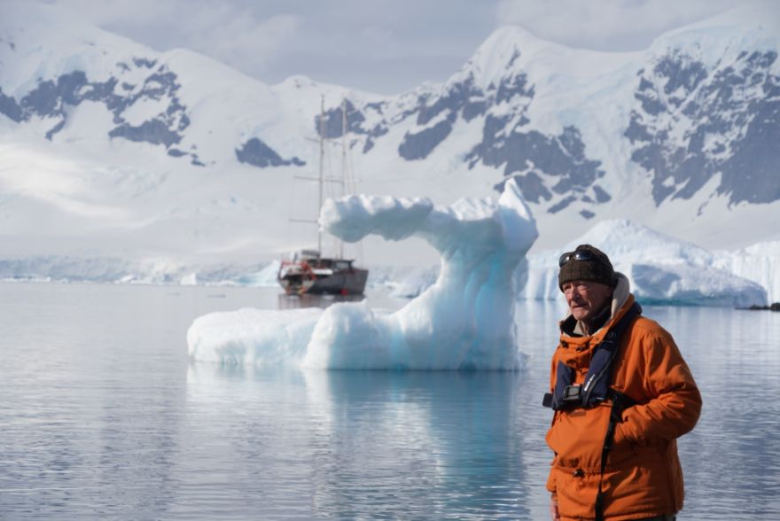 Man in an orange parka stands at a chilly shore with a blue iceberg arch and a sailing ship in the background.