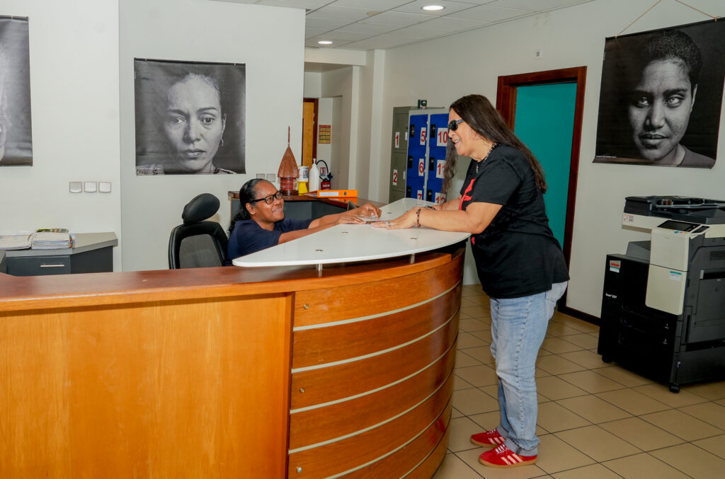 2026-04-05 association valentin haüy 2 • province sud Woman at a curved wooden reception desk hands documents to a seated staff member behind the counter in a lobby area with large portraits on the walls