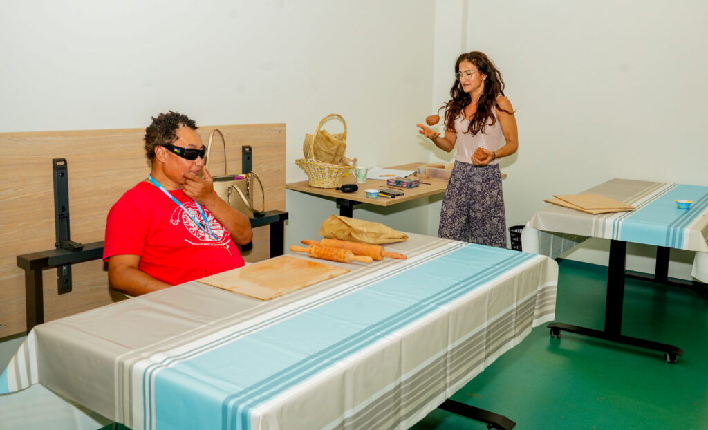 2026-04-05 association valentin haüy 3 • province sud Man in a red shirt with sunglasses sits at a long table as a woman in a light top tosses a potato in the air in a room with food and bags nearby