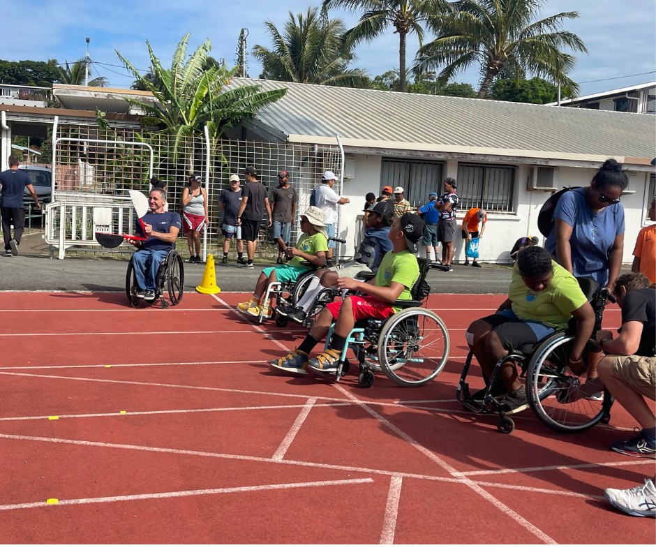 Group of people in wheelchairs on a sunny outdoor track with spectators and palm trees in the background