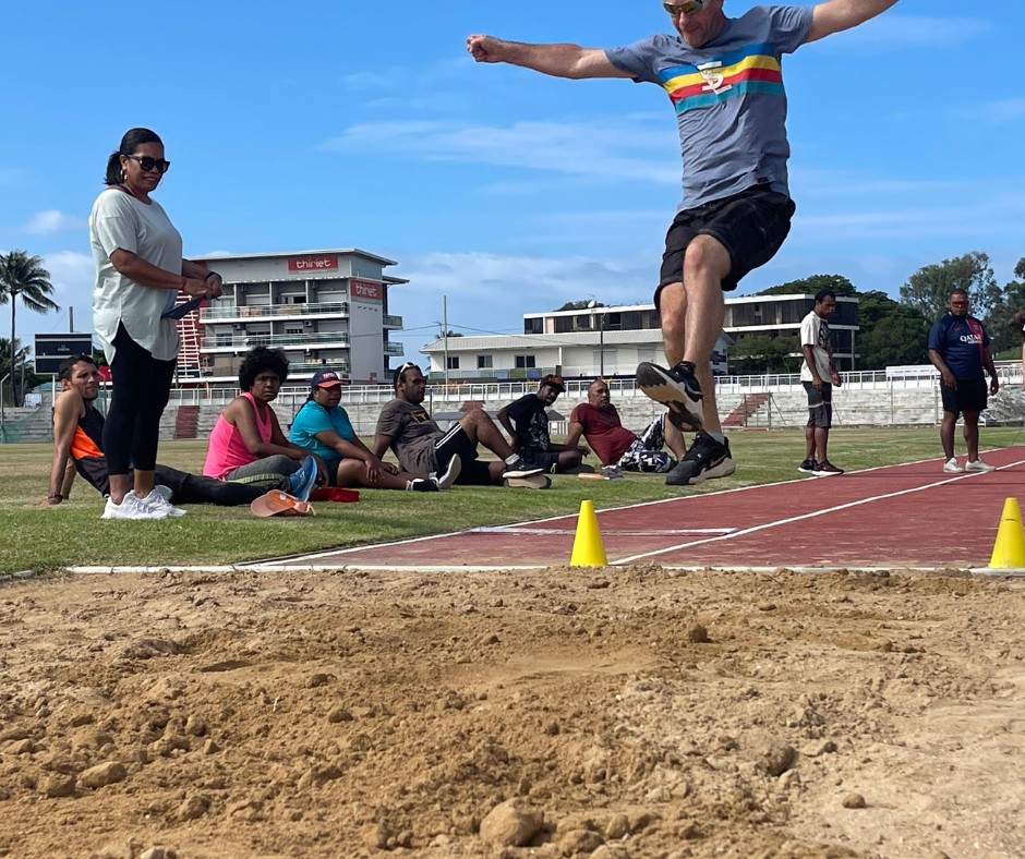 Athlete mid air during a long jump over the sand pit as teammates watch on a sunny track field