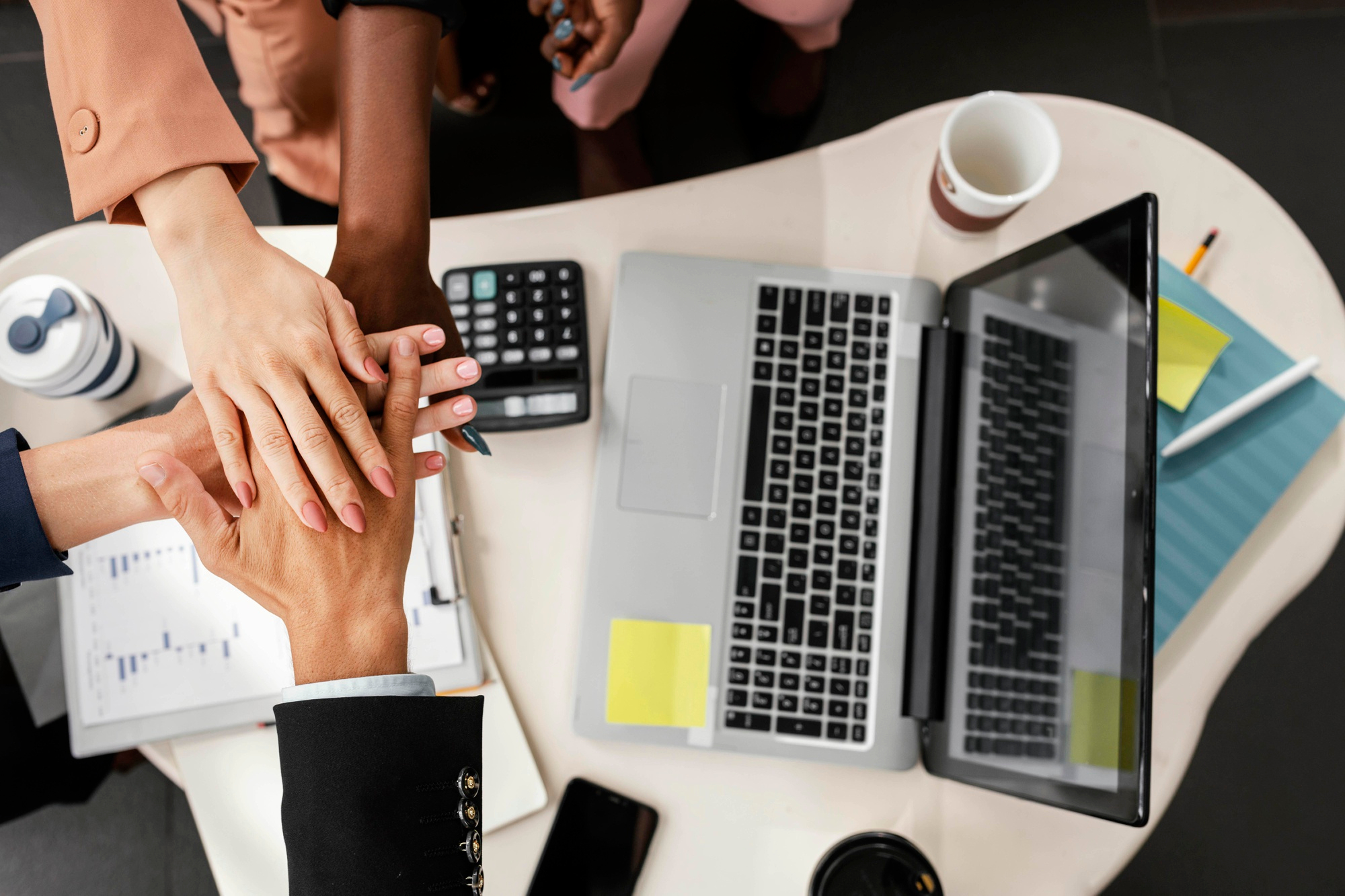 Team members stack hands in a circle over a desk with laptops, papers, and coffee cups, showing teamwork in an office.