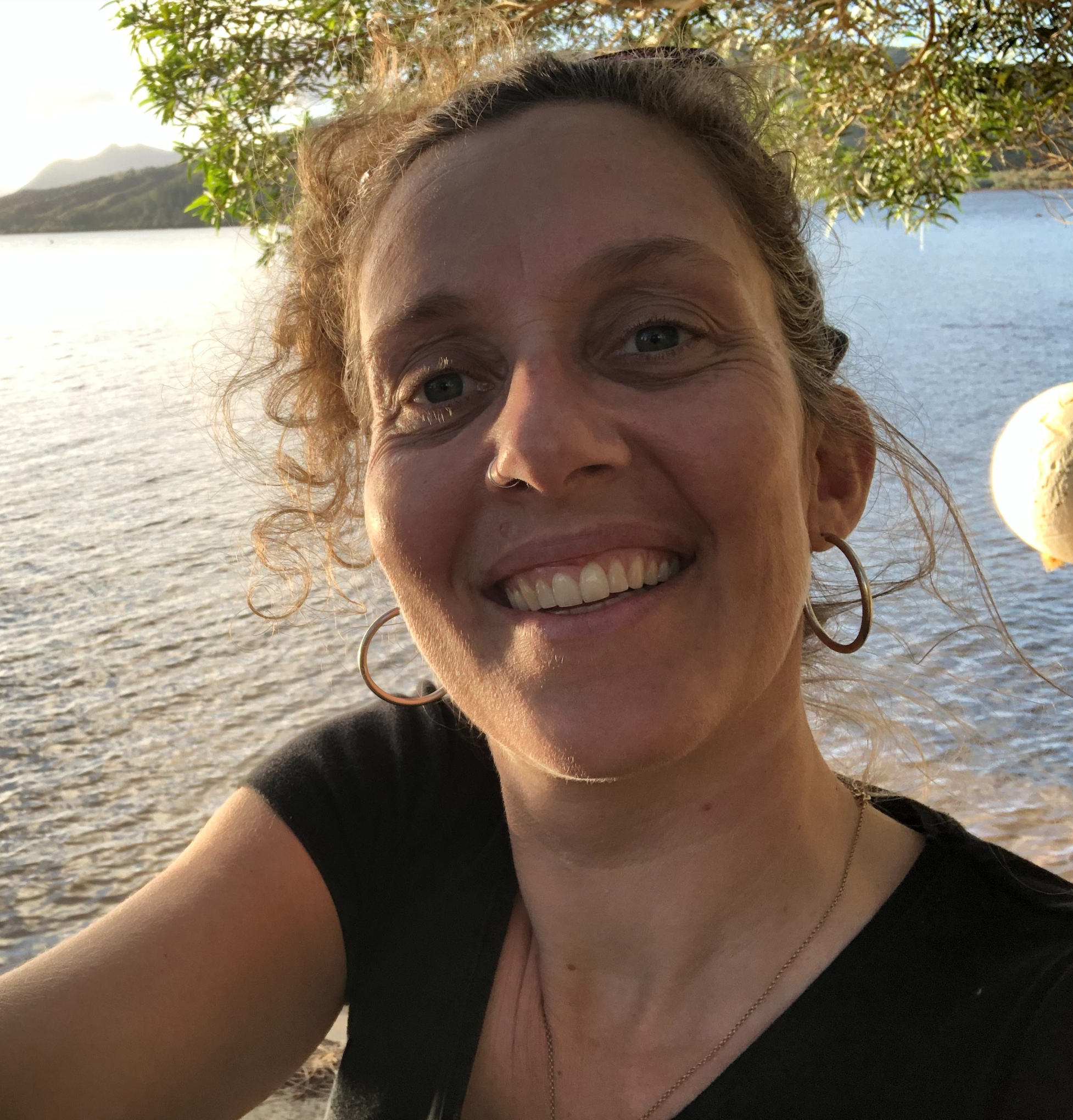 Close-up of a smiling woman in a black top by a lake, with trees and hills in the background.