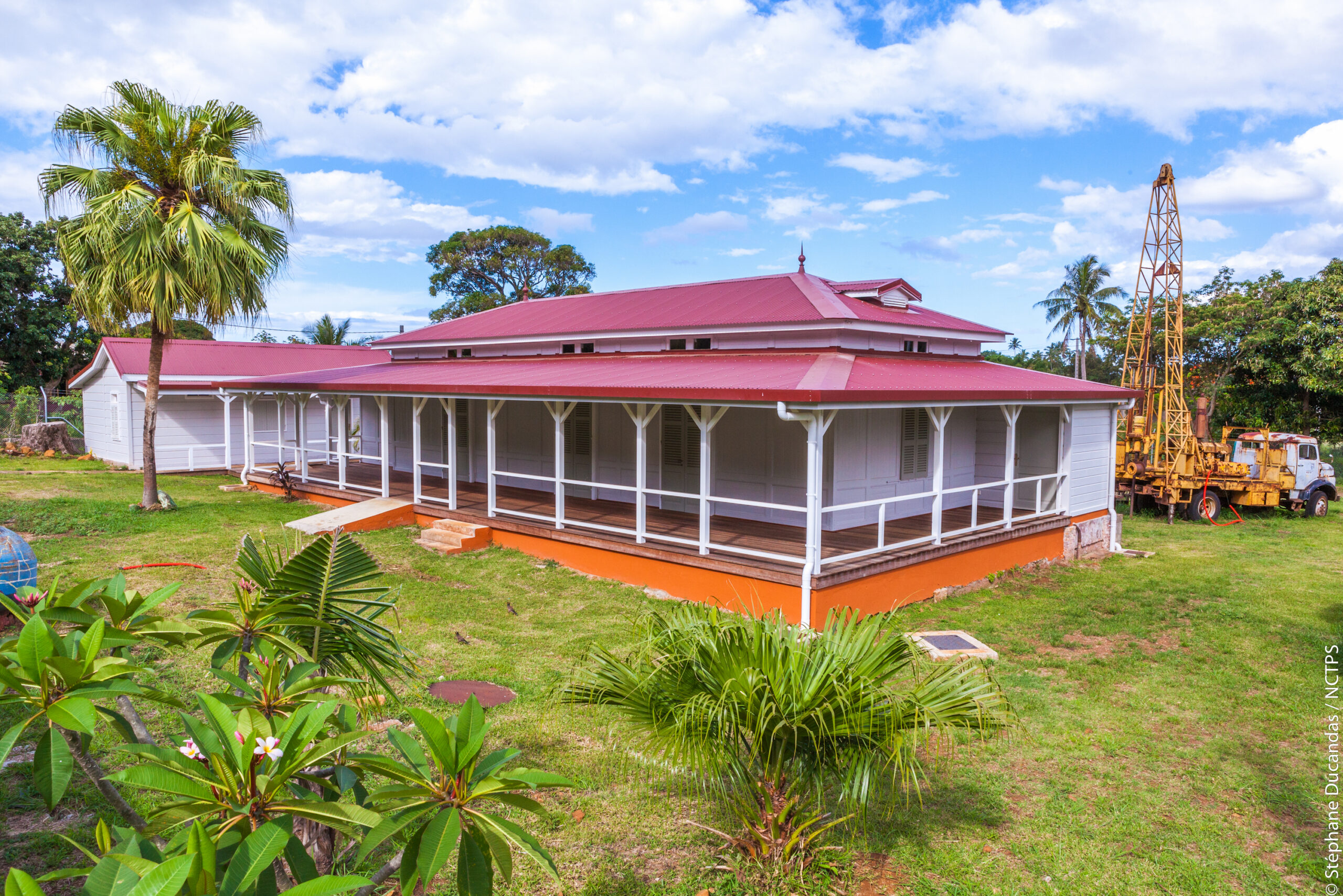 White house with a red metal roof and wraparound porch, set in a tropical yard with palm trees and a blue sky.