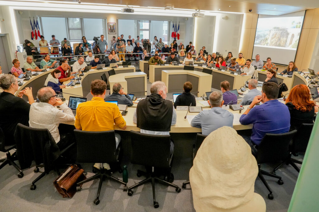 2 • province sud Council meeting in progress many people seated around a curved multitiered desk with laptops while an audience watches from the back of the room under flags and a projector screen