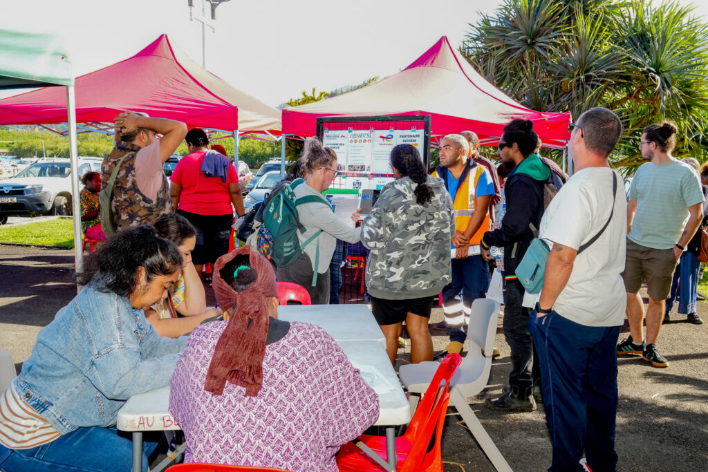3 • province sud People gather around a booth under red canopy tents at an outdoor community event with a display board and people waiting in line