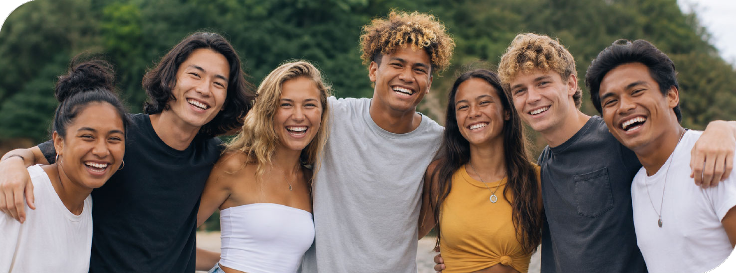 Seven diverse friends with arms around each other, smiling outdoors.