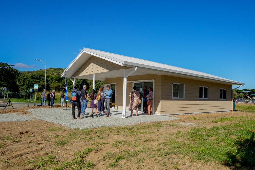 A group of people gathers outside a single story beige building with a white slanted roof on a sunny day