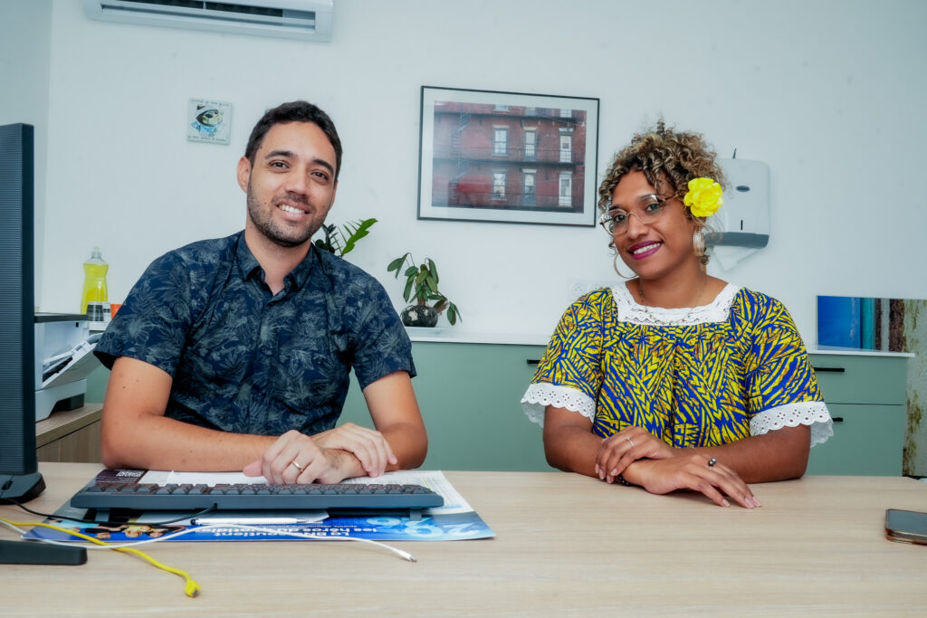 Dsc08047 • province sud Two colleagues sit at a desk in an office smiling at the camera a man on the left and a woman with a yellow flower in her hair on the right