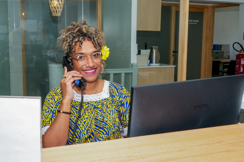 Dsc08063 • province sud Smiling woman wearing glasses and a yellow floral dress talks on a landline phone at a wooden reception desk in an office setting