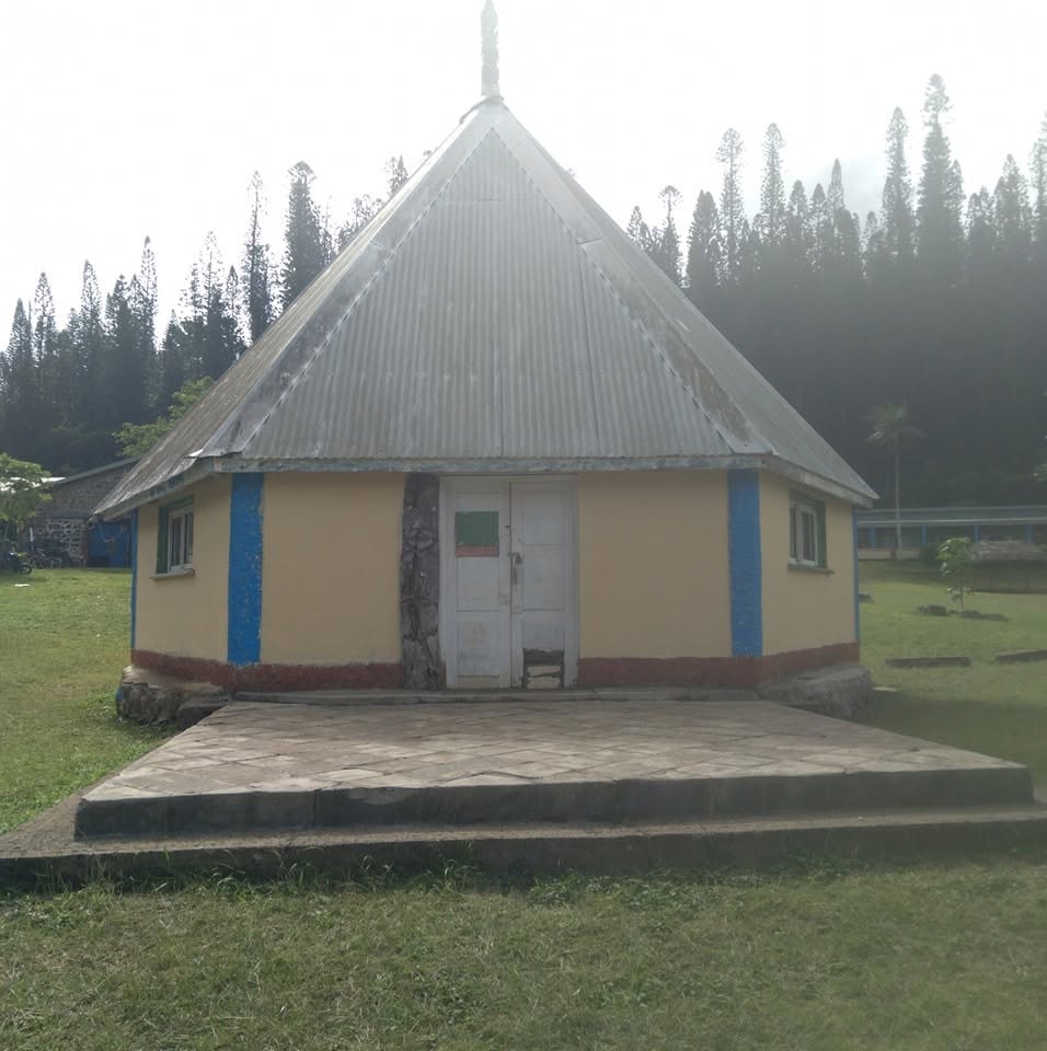 Small yellow hut with a tall, gray metal roof and blue trim, sitting on a raised stone patio in a grassy area.