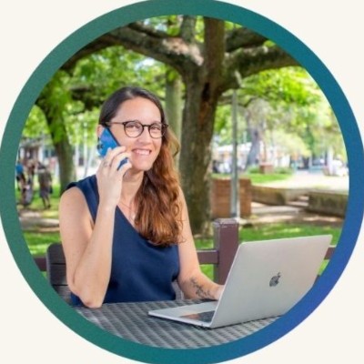 Woman seated at an outdoor cafe table, talking on a smartphone while using a silver laptop.