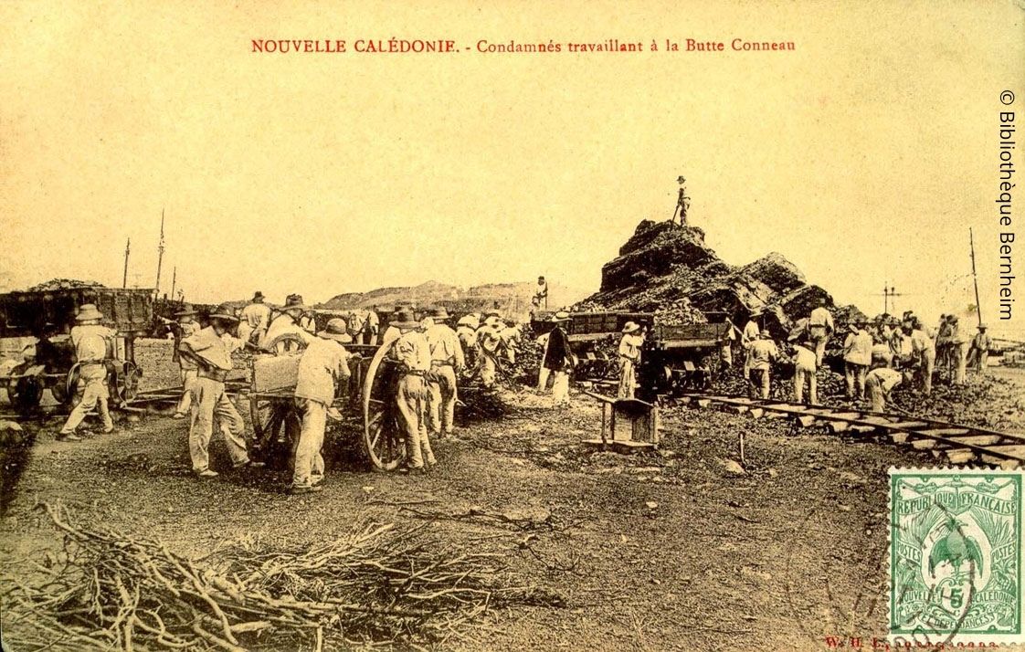Historical postcard of a mining site in New Caledonia with workers loading ore around a steam cart and a large pile of rocks in the background.