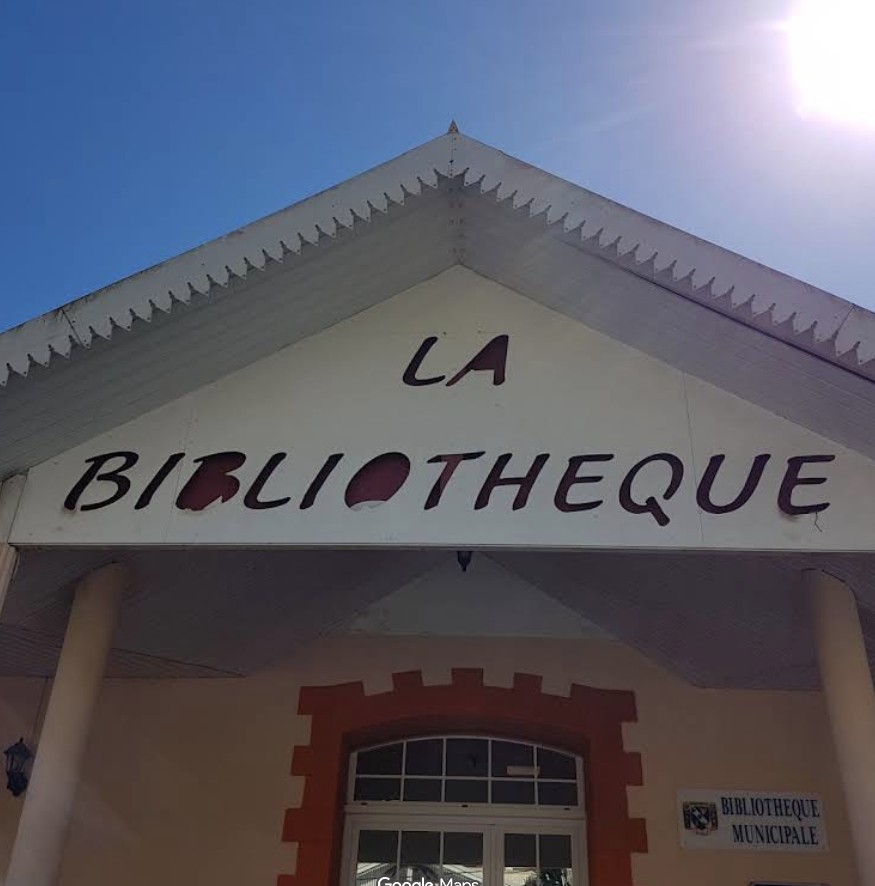 Front of a municipal library building with a triangular roof and a large sign reading 'LA BIBLIOTHEQUE' on the facade, under a clear blue sky.