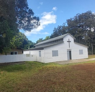 White wooden church with a cross on the front, set in a grassy yard under a blue sky with trees nearby.