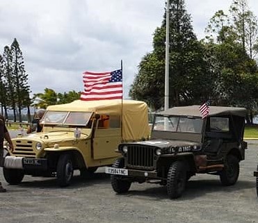 Two vintage military jeeps with American flags parked side by side on a paved lot under a cloudy sky, beige and dark green models.