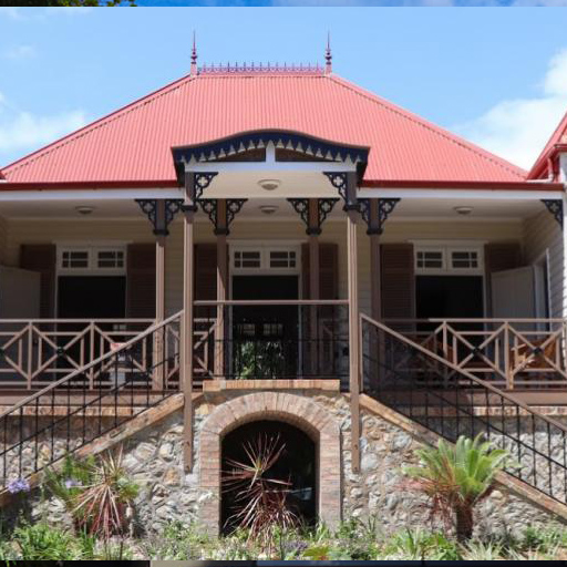 Two-story house with a red metal roof, decorative porch supports, and a stone foundation with stairs leading to a central entrance.