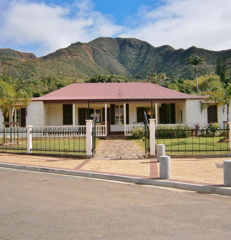 Single-story white house with a red metal roof, a wrought-iron fence, and a stone walkway leading to the front door, set against green lawn and mountains in the background.