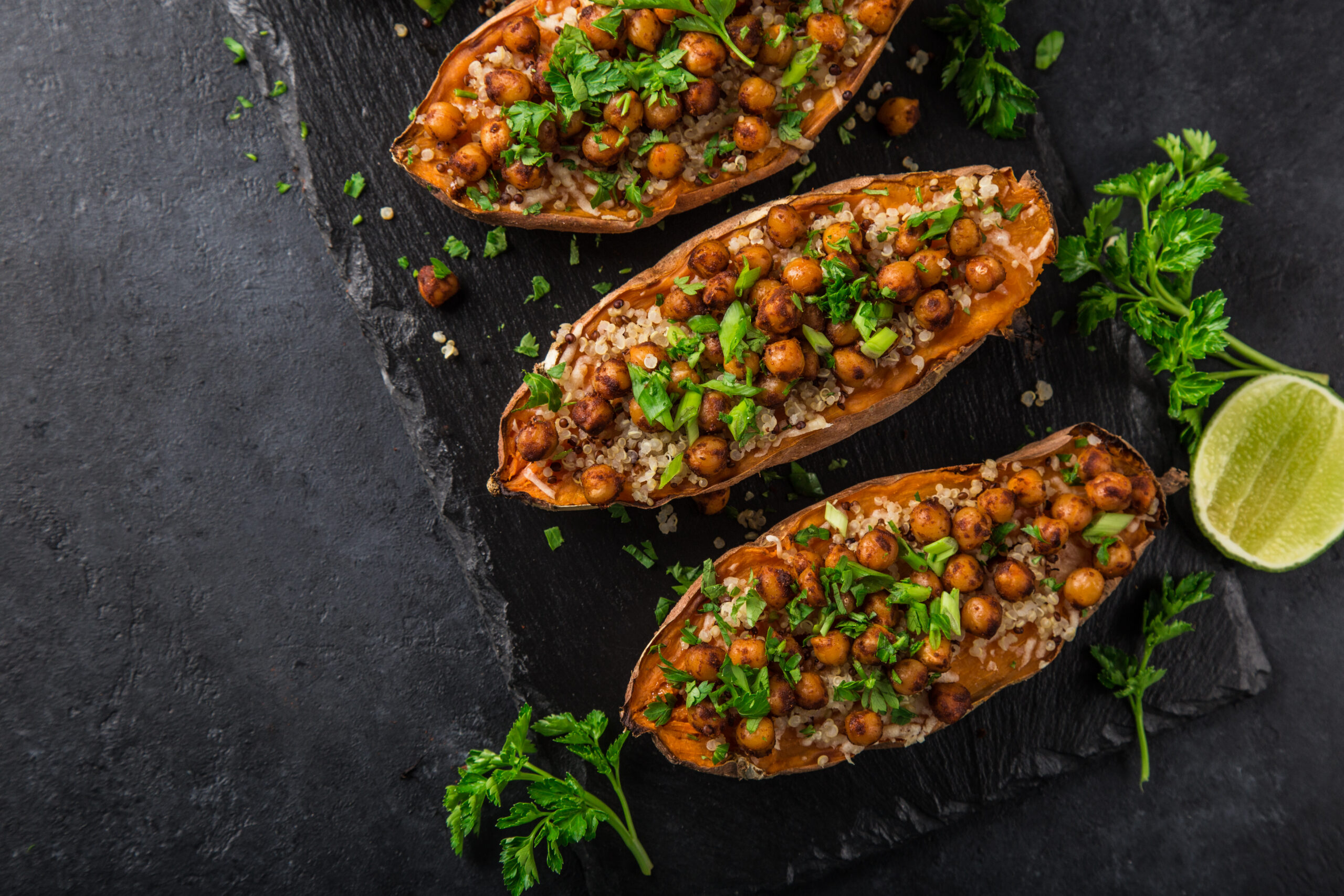 Roasted sweet potato halves stuffed with quinoa, chickpeas, and chopped parsley on a dark slate surface with lime wedges nearby.