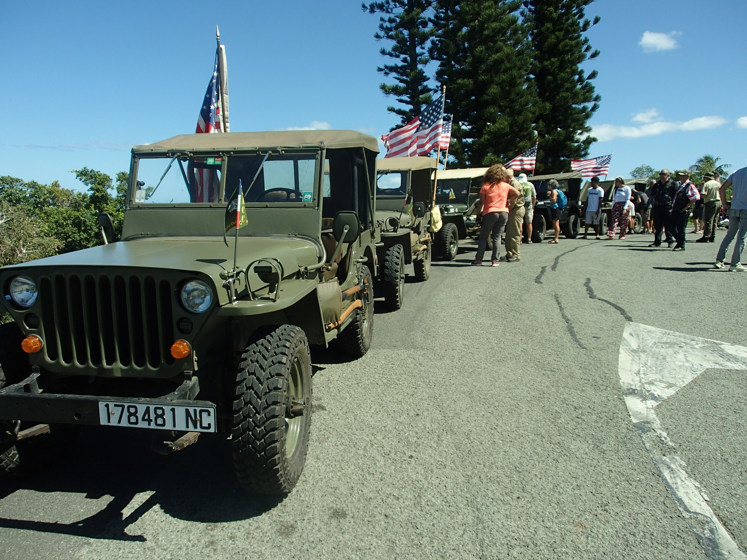 Line of olive drab military jeeps with American flags along a sunny road, spectators gathered in the background under a blue sky.
