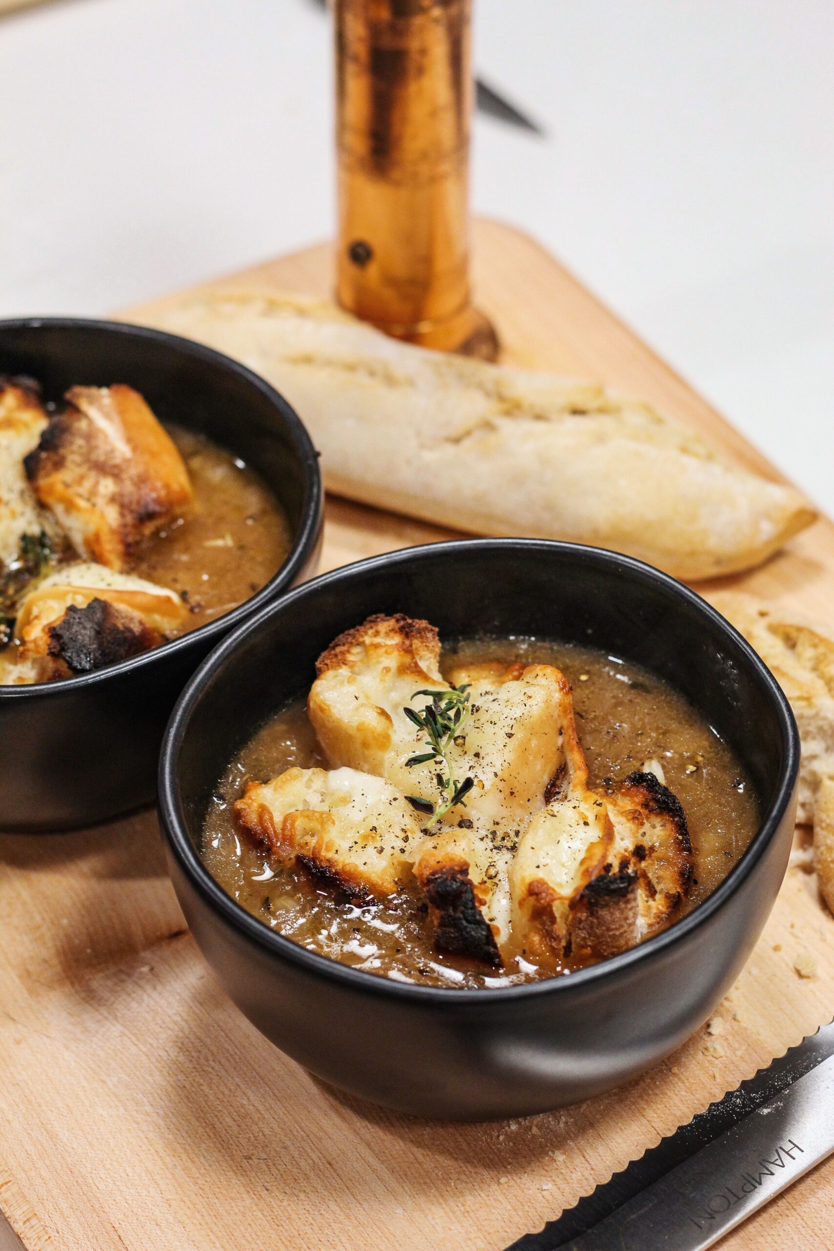 Two black bowls of creamy soup topped with croutons and herbs on a wooden board, with a torn baguette in the background.