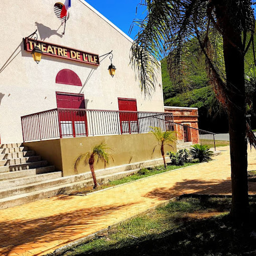 Exterior of Theatre de l'Île with red doors, balcony railing, and palm trees on a sunny day by a brick walkway