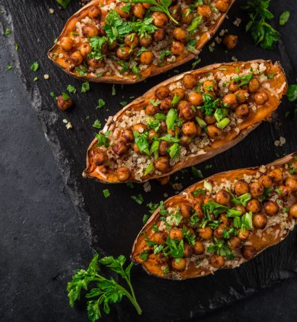 Roasted sweet potato halves stuffed with quinoa, chickpeas, and chopped parsley on a dark slate surface with lime wedges nearby.