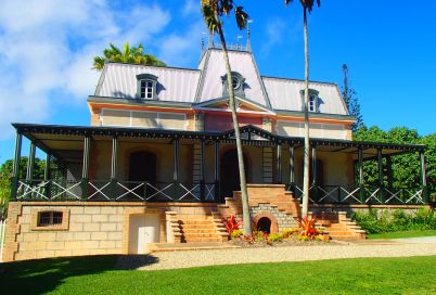 Two-story house with a metal roof, brick steps, and a wraparound veranda under a bright blue sky with palm trees in front.