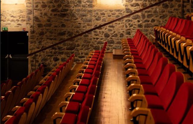 Empty theater with red upholstered seats and yellow armrests arranged in rows along a stone wall.