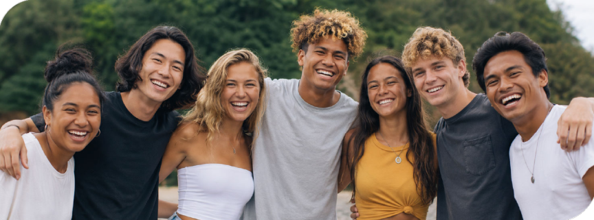 Seven diverse friends with arms around each other, smiling outdoors.