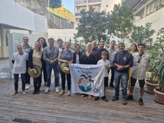 Group of diverse people posing outdoors on a wooden deck, holding a banner that says 'Ambassadeur du lagon' and smiling at the camera.
