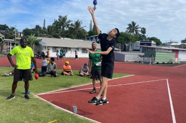 Two athletes leap to strike a blue ball during a track-side sport drill while teammates watch on a sunny day.