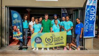 Group of diverse volunteers in turquoise shirts pose together behind a bright yellow banner at an outdoor event doorway, with Province Sud banners nearby.