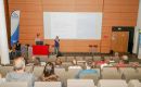 Speaker at a red lectern presenting to an audience in a conference hall, with a large projected slide on a wood-paneled wall behind them.