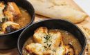 Two black bowls of creamy soup topped with croutons and herbs on a wooden board, with a torn baguette in the background.