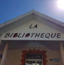 Front of a municipal library building with a triangular roof and a large sign reading 'LA BIBLIOTHEQUE' on the facade, under a clear blue sky.