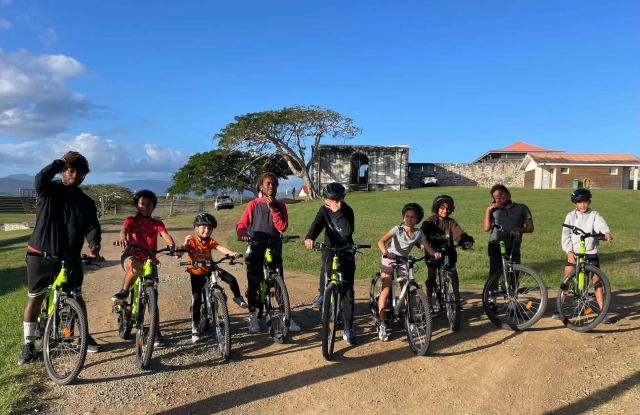 Group of kids on bright green bikes with helmets, posing on a sunny dirt path in a park with a grassy field behind.