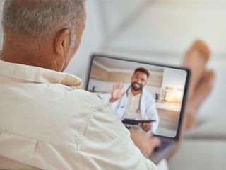 Older man shown from behind holding a tablet during a telemedicine video call, doctor waving on screen