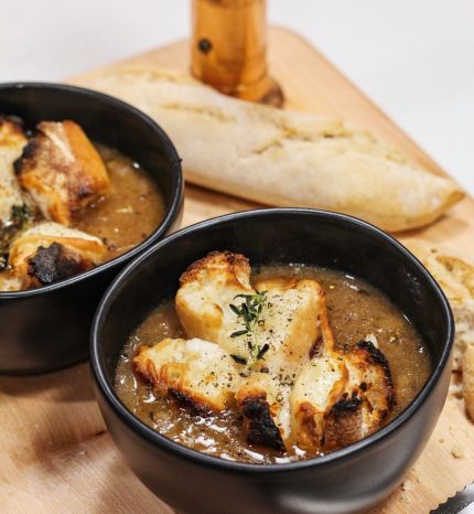 Two black bowls of creamy soup topped with croutons and herbs on a wooden board, with a torn baguette in the background.