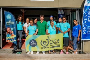 Group of diverse volunteers in turquoise shirts pose together behind a bright yellow banner at an outdoor event doorway, with Province Sud banners nearby.