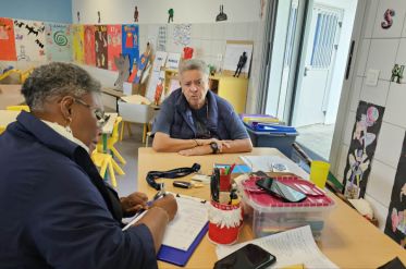 Two adults sit at a table in a bright art room, with colorful student artworks on the walls.