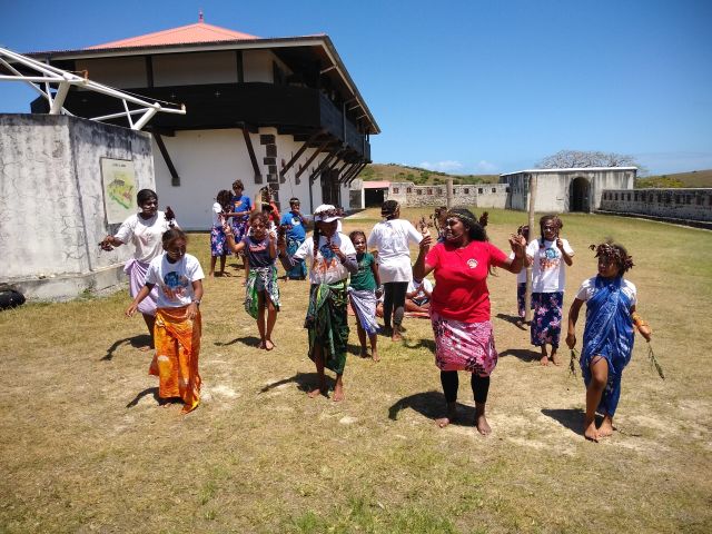 Group of children in bright sarongs dancing outdoors in a historic fort complex under a clear blue sky.