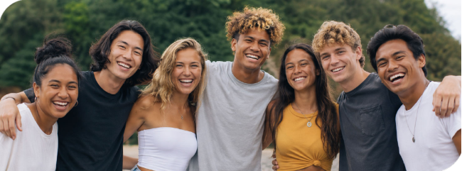 Seven diverse friends with arms around each other, smiling outdoors.