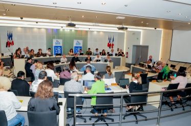 Large conference room with a panel of speakers at the front, French flags behind them, and attendees using laptops and notebooks.