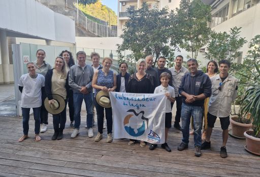 Group of diverse people posing outdoors on a wooden deck, holding a banner that says 'Ambassadeur du lagon' and smiling at the camera.