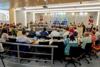 Panel of officials seated at a long desks in a conference room, with an audience using laptops and microphones; flags and banners in the background. Council meeting in progress.