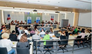 Large conference room with a panel of speakers at the front, French flags behind them, and attendees using laptops and notebooks.